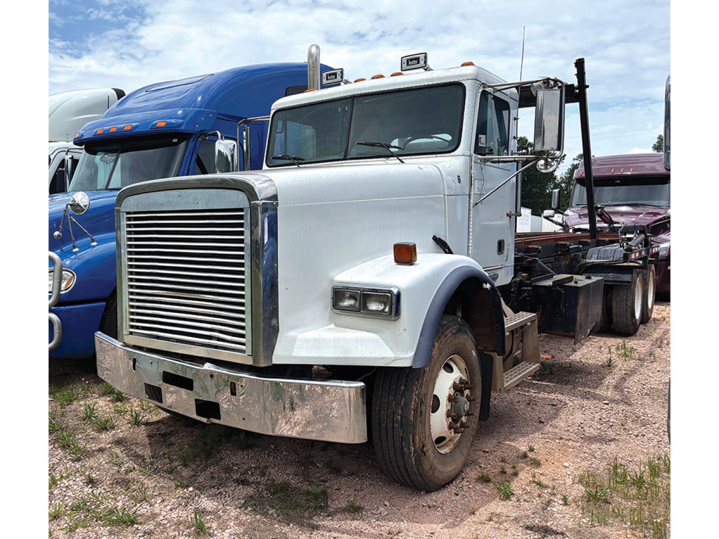 Front-left view of a white Freightliner FLD120SD truck in a lot, with a large grille, bumper, and dual rear axles, other trucks in background.