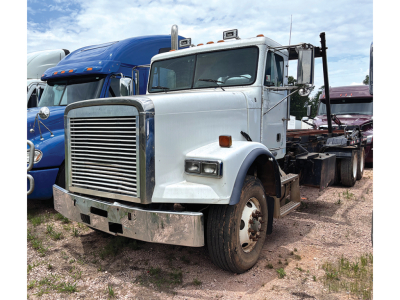 Front-left view of a white Freightliner FLD120SD truck in a lot, with a large grille, bumper, and dual rear axles, other trucks in background.
