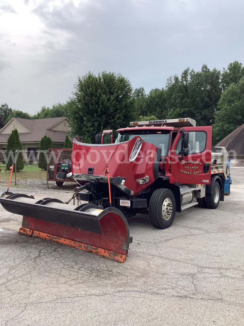 Truck showing front-mounted snow plow and red Freightliner 108SD dump truck in a parking lot.