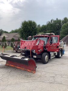 Truck showing front-mounted snow plow and red Freightliner 108SD dump truck in a parking lot.