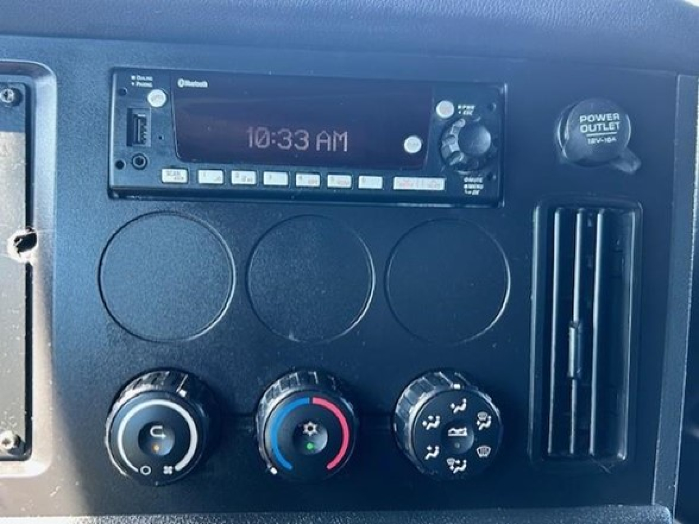 Dashboard panel of a Freightliner Cascadia shows radio unit with time 10:33 AM, three circular gauges, climate control knobs, and a power outlet; manual knobs and vents visible.