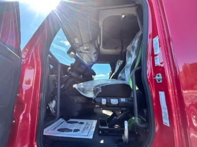 Cabin interior of a red Freightliner Cascadia 126 from the driver side door open, showing seat with plastic cover, steering wheel, and dashboard area.