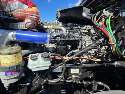 Engine compartment of a Freightliner Cascadia showing top view with blue intercooler hose, multiple wiring harnesses, fluid reservoirs, and engine components.