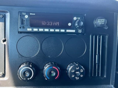 Dashboard panel of a Freightliner Cascadia shows radio unit with time 10:33 AM, three circular gauges, climate control knobs, and a power outlet; manual knobs and vents visible.