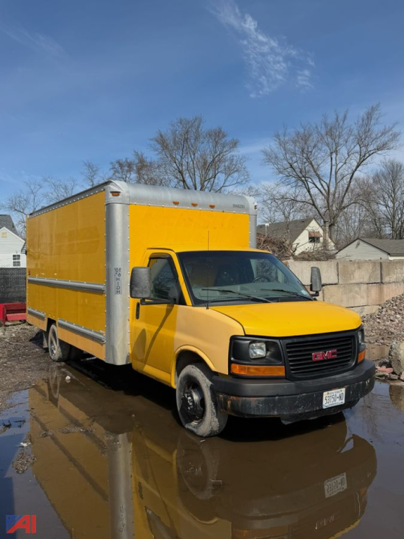 GMC Savana box truck in yellow with a large cargo box and cab, front view, parked on wet ground.