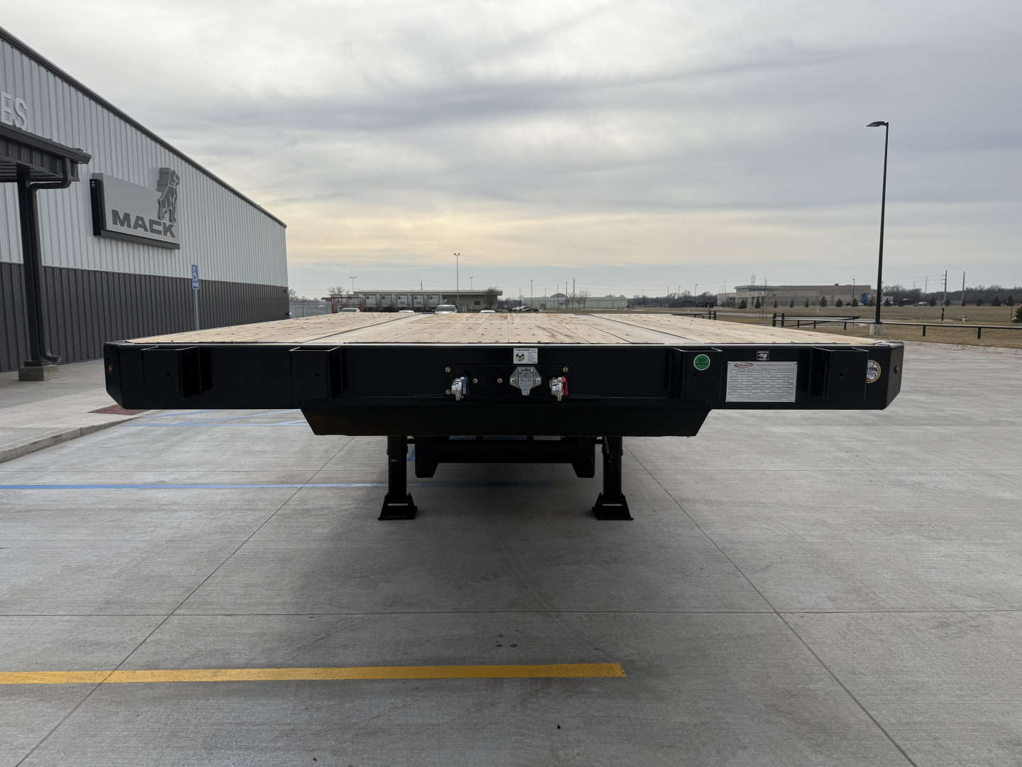 Flatbed trailer rear view with deck and landing gear, showing hydraulic connections and safety labels.