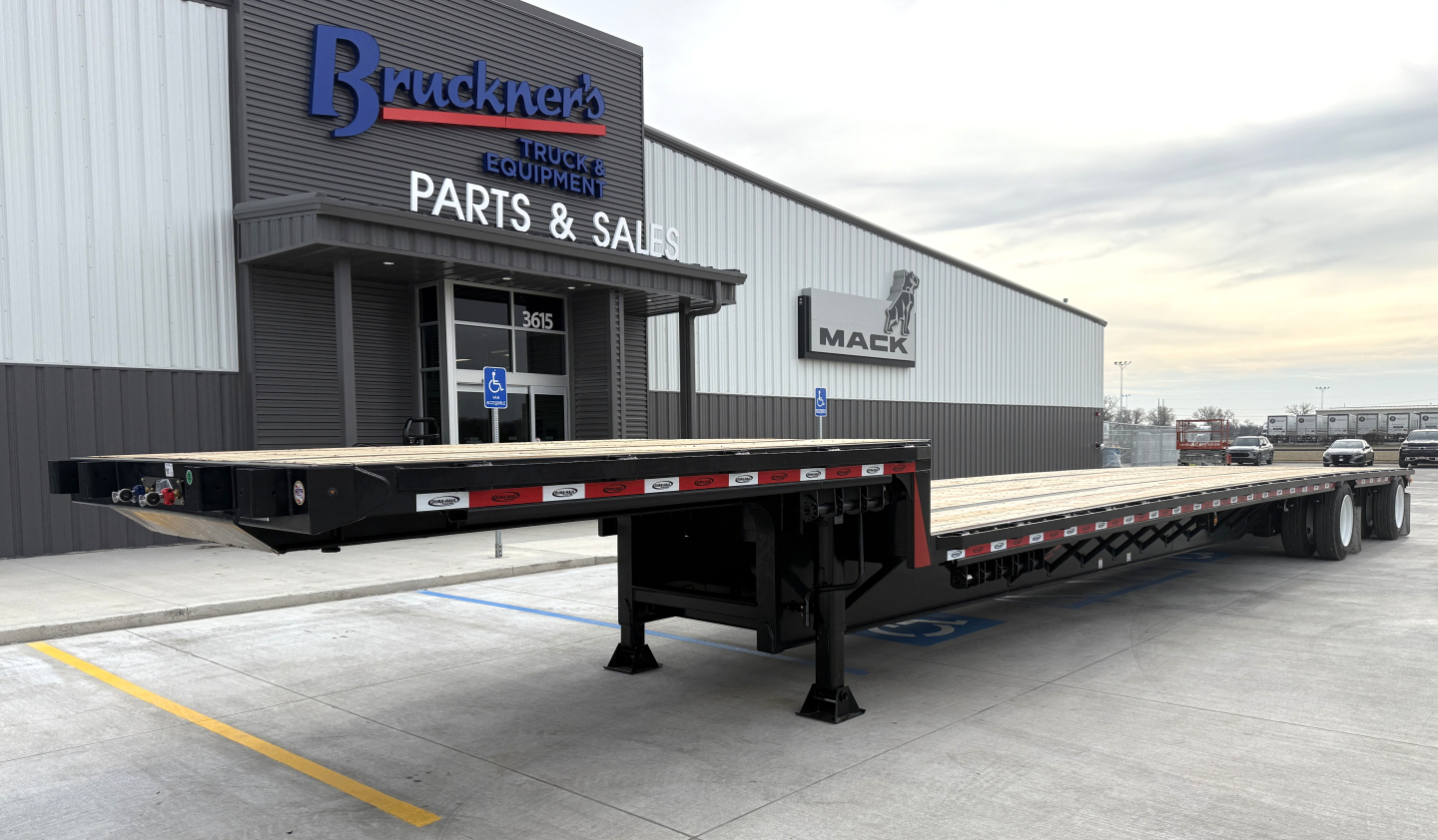 XX trailer long flatbed drop deck showing black steel frame, extended gooseneck, and tandem axles in a dealership lot.