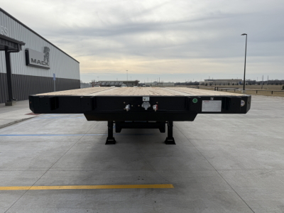 Flatbed trailer rear view with deck and landing gear, showing hydraulic connections and safety labels.