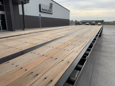 Flatbed trailer deck showing wooden planks and steel frame near Mack building, urban lot in background.