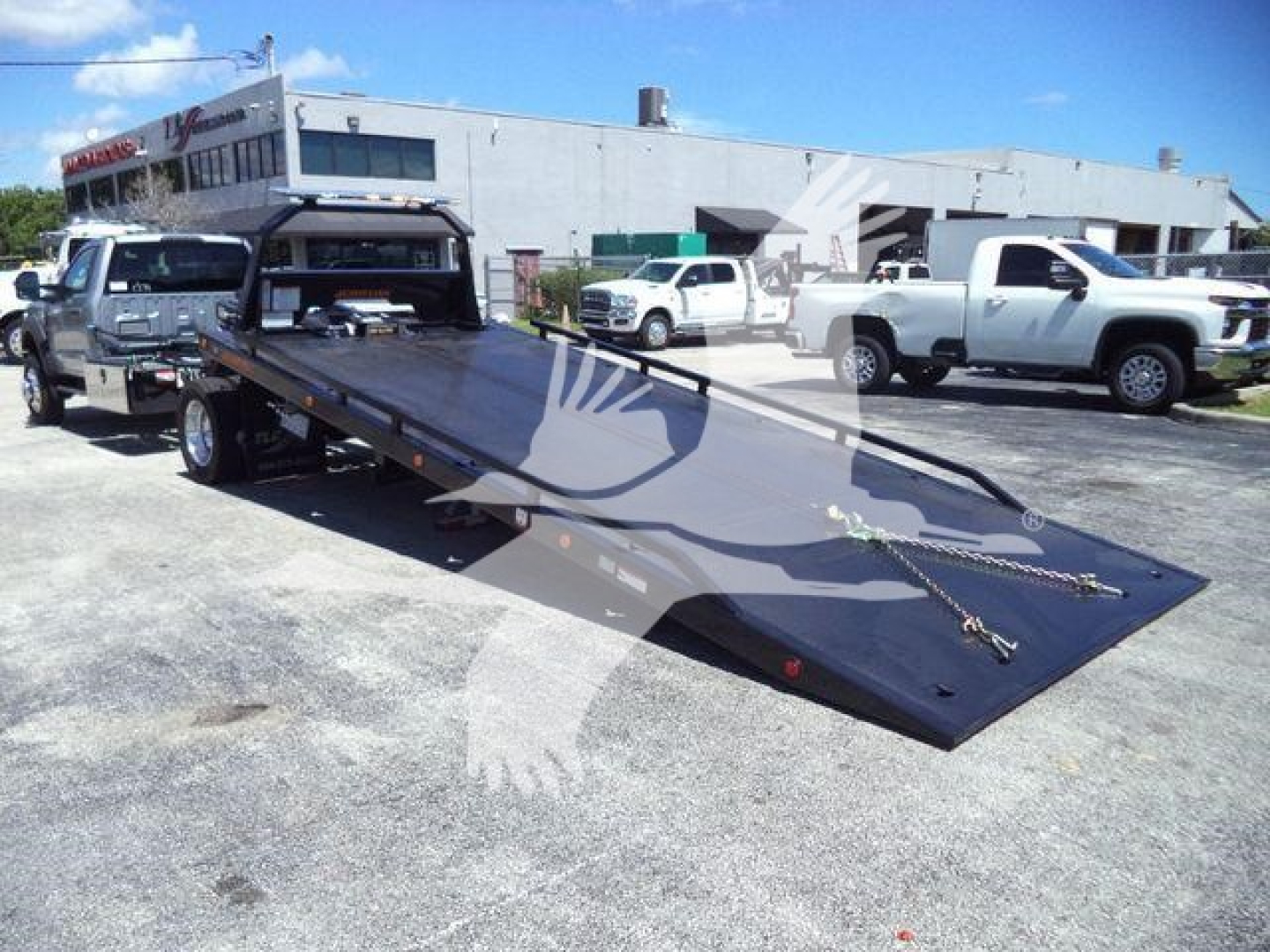 Flatbed tow truck with long ramp in a dealership lot, Ford F-Series chassis visible, multiple trucks in background.