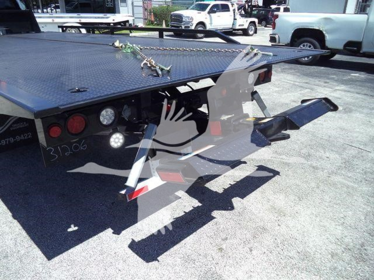 Flatbed rollback tow truck rear with ramp and chains, flat black steel deck, rear lights and reflective markers visible.