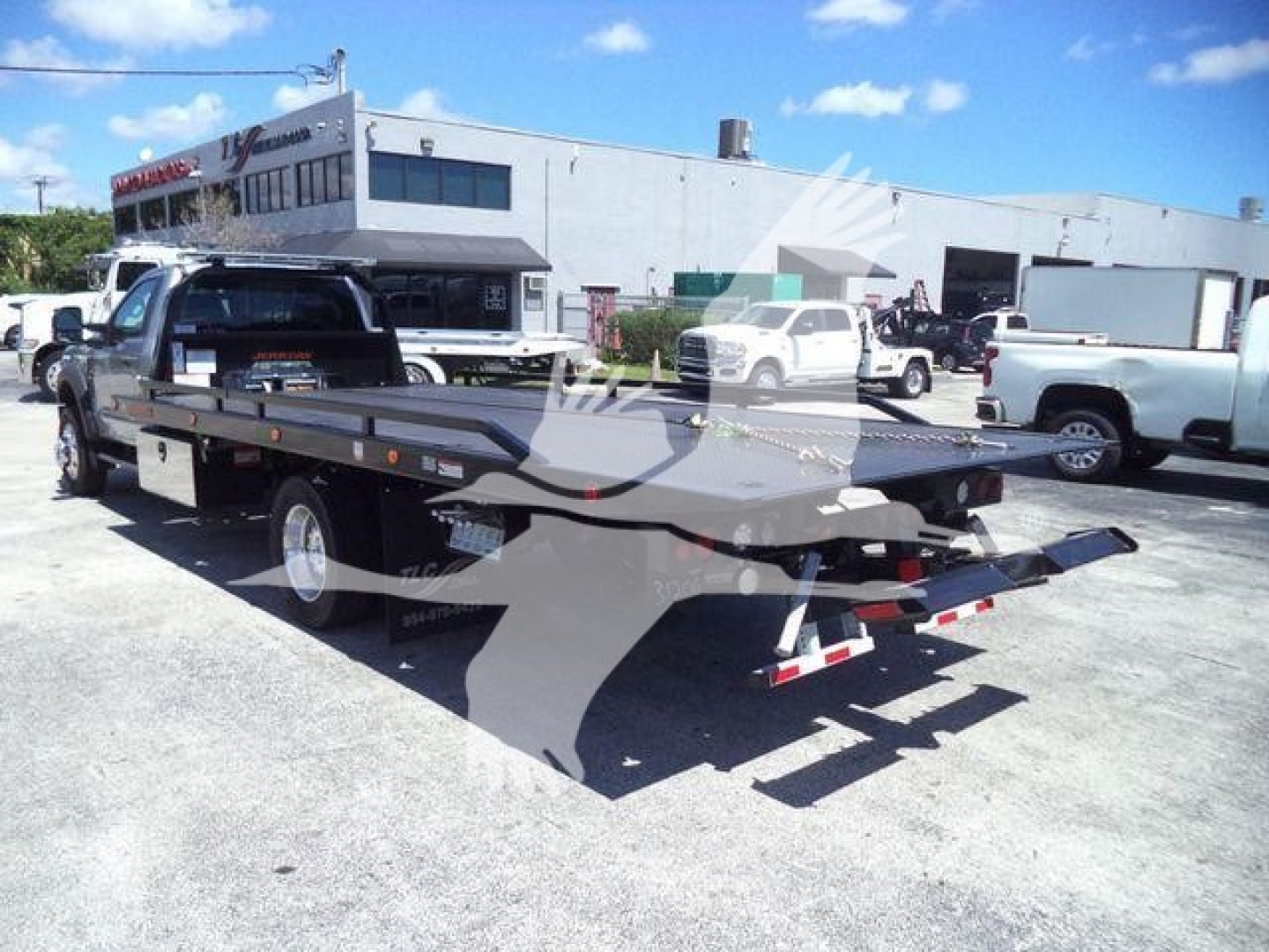 Flatbed rollback tow truck with extended flatbed and rear ramps visible, parked in lot with building and other trucks in background.
