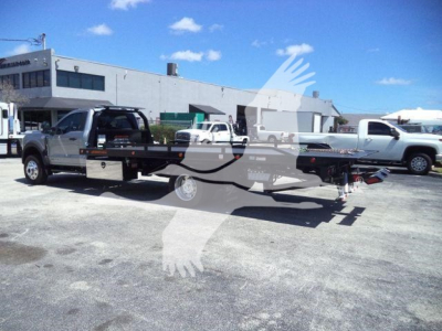Flatbed tow truck on lot with other vehicles; large building in background; blue sky.