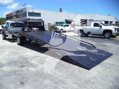 Flatbed tow truck with long ramp in a dealership lot, Ford F-Series chassis visible, multiple trucks in background.