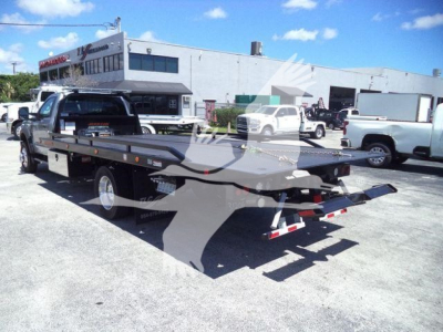 Flatbed rollback tow truck with extended flatbed and rear ramps visible, parked in lot with building and other trucks in background.