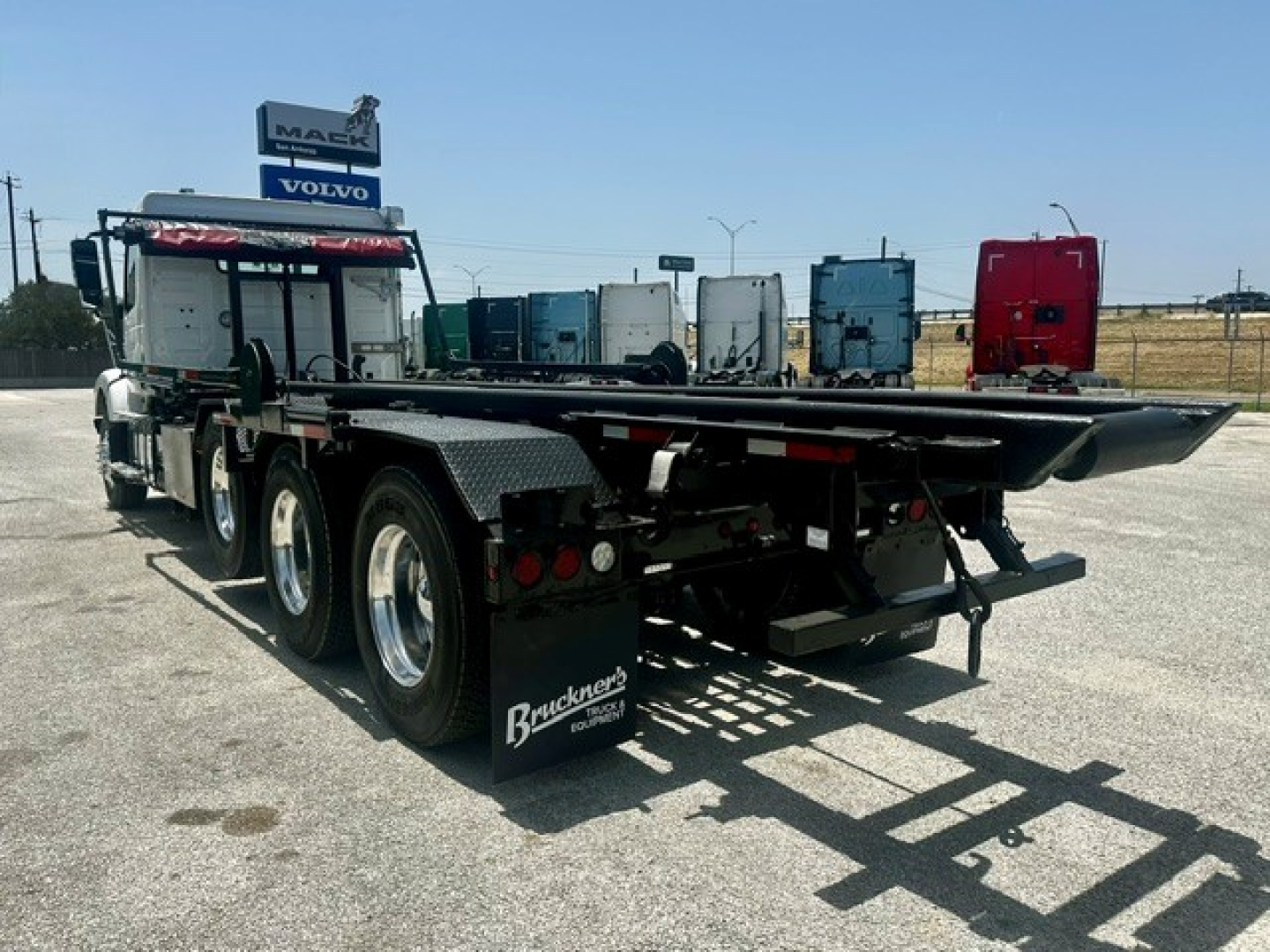 Flatbed trailer attached to a Volvo VHD truck in a dealership lot, with multiple semi trucks in background.