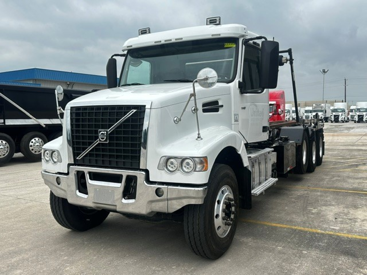 Volvo VHD rolloff truck in white, front-left view with grille, headlights, chrome bumper, and side mirrors.