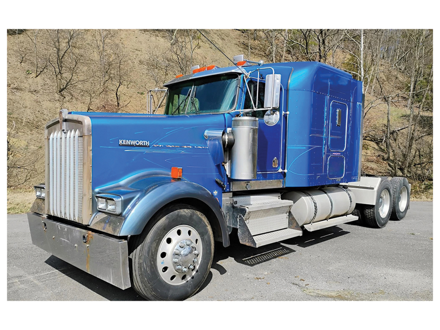 Kenworth W900L heavy-duty truck front-left view showing blue sleeper cab and chrome fuel tanks.