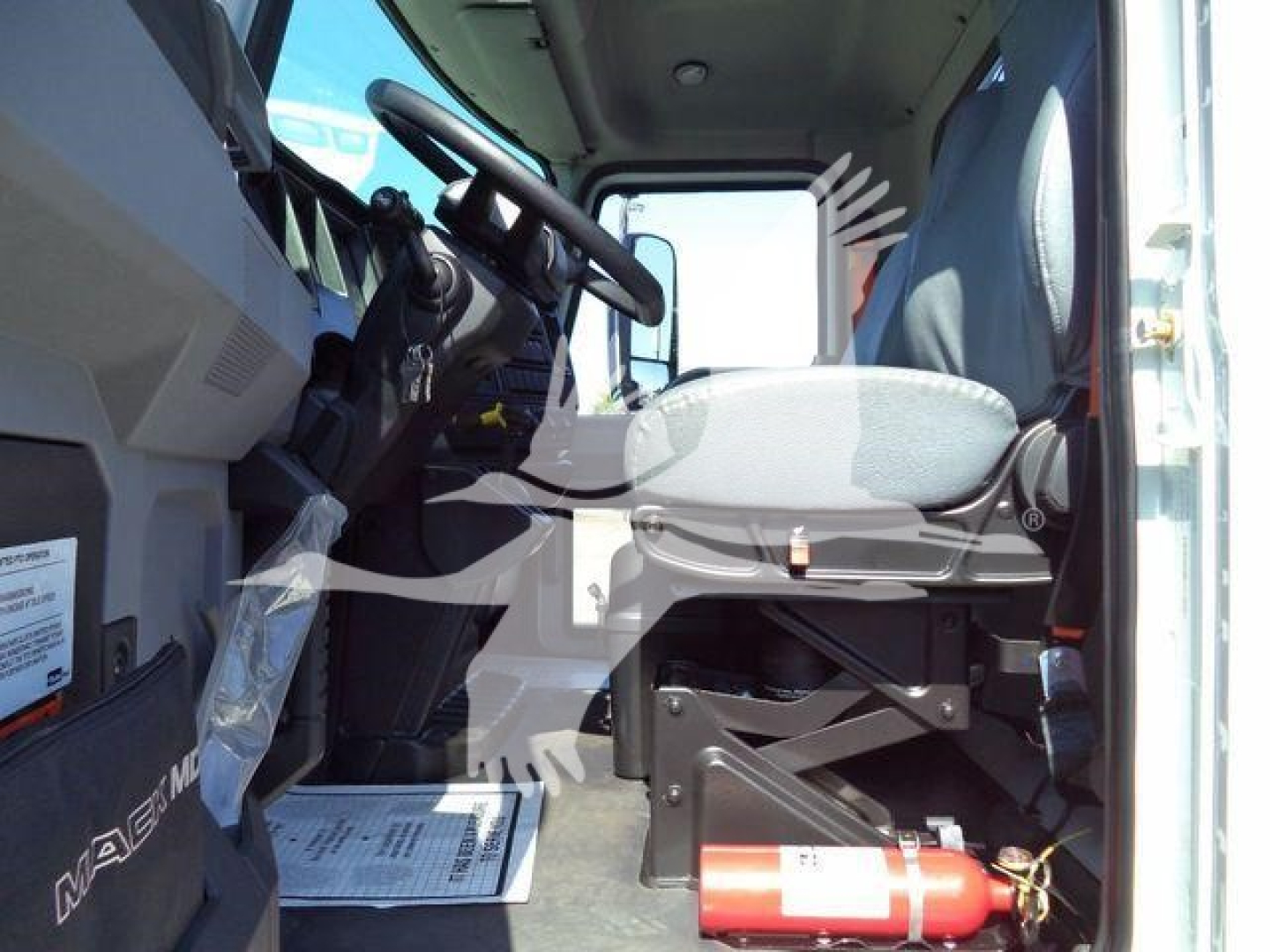 Cab interior of a 2026 Mack MD6 showing driver seat, steering wheel, and dashboard; fire extinguisher mounted under the seat area; view from open door.