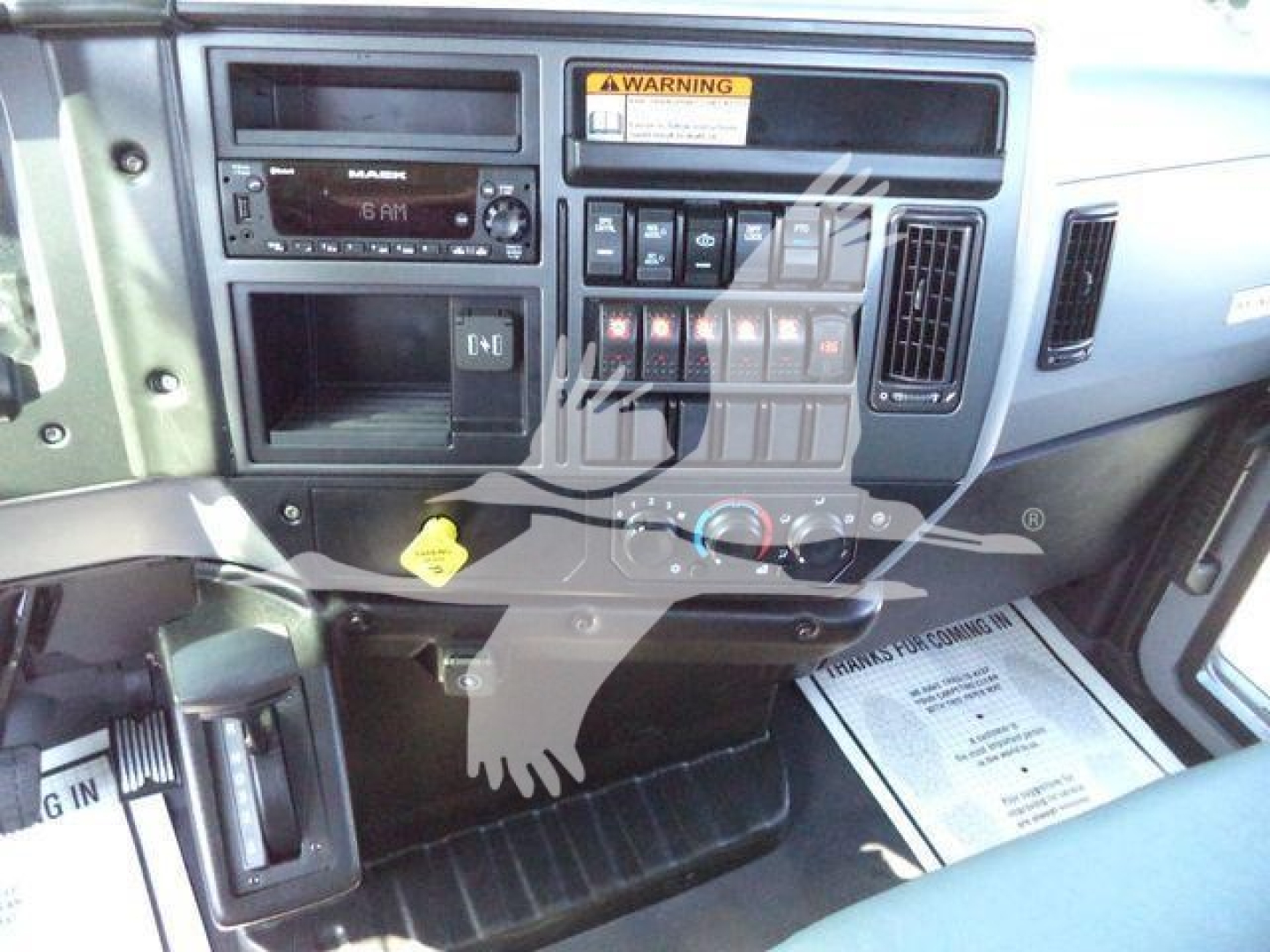 MD6 Mack truck interior showing dashboard controls, radio, switches, and warning label. Center console with climate controls and multiple toggle switches. Steering area with pedal and floor mat visible.
