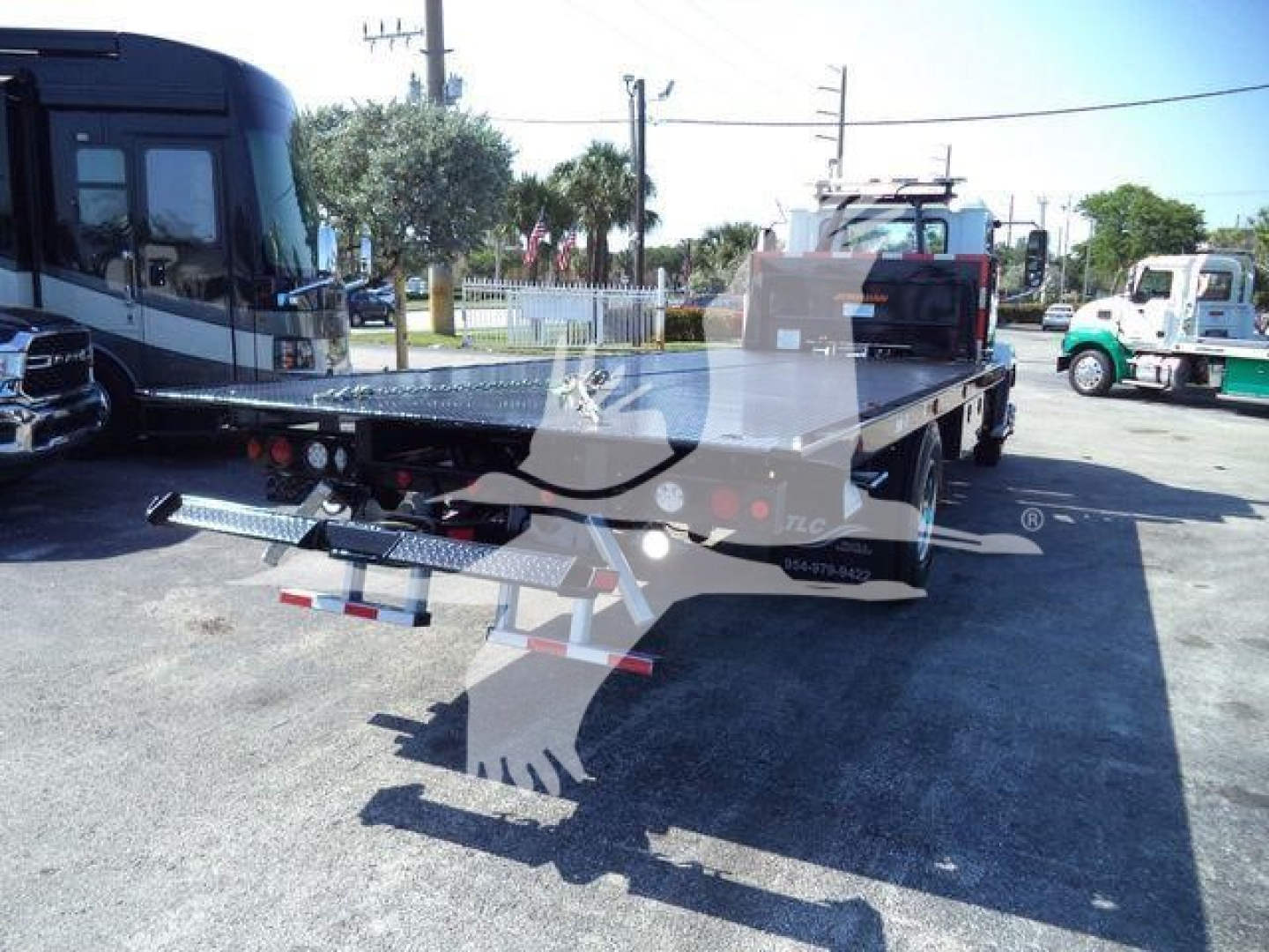 Flatbed tow truck/dolly with rear ramps and vehicle wheel-lift area, Mack MD6 yard unit in background, showing tow equipment and platform.