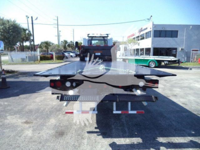Flatbed tow truck trailer viewed from rear showing reflective tape and taillights; Mack MD6 rollout truck in background.