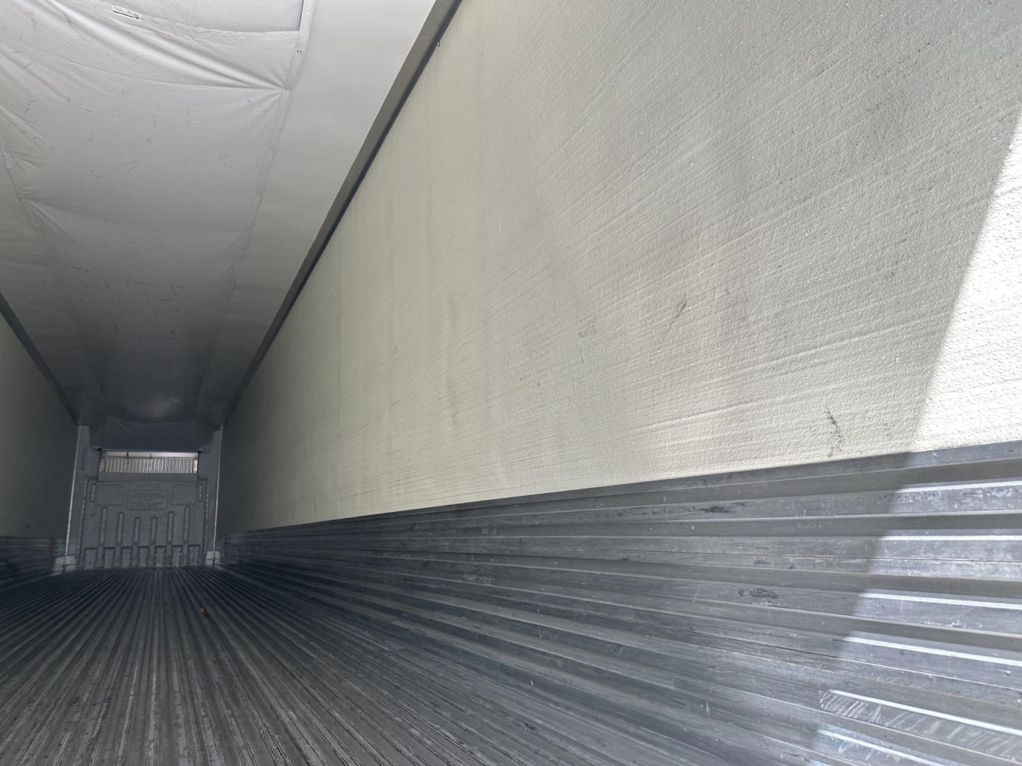Interior of a trailer cargo area showing ribbed metal side walls and floor with a rear door gap; no legible labels or fluid reservoirs visible.