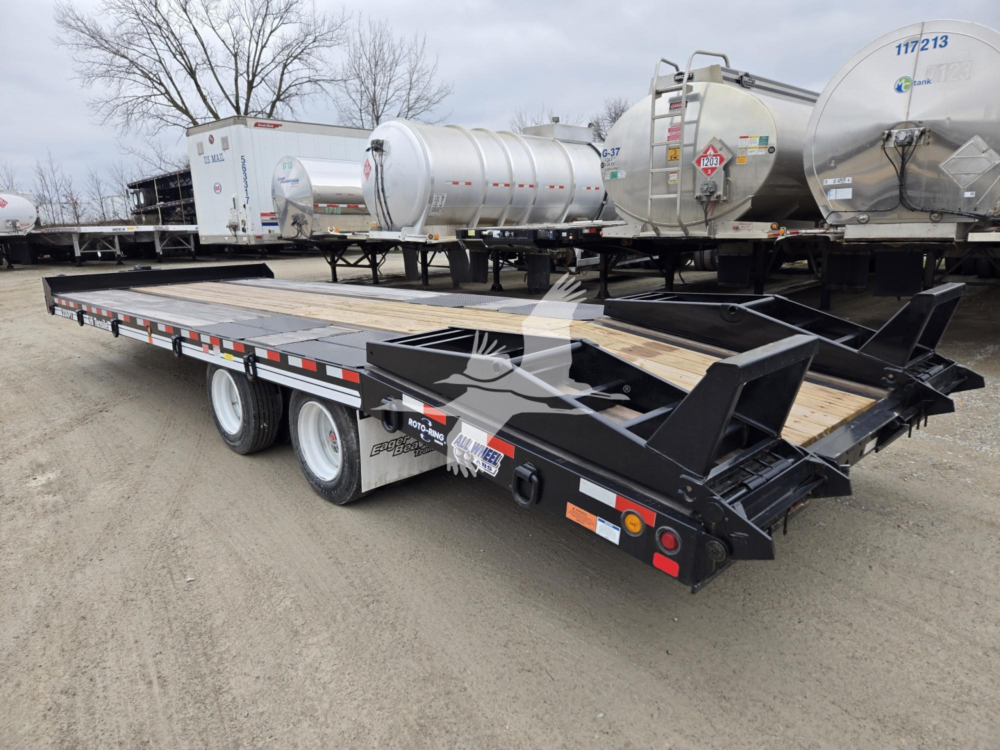 Trailer bed with dual rear axles and metal framework showing a flatbed trailer for transporting tanks near other trailers and stainless tanker units.