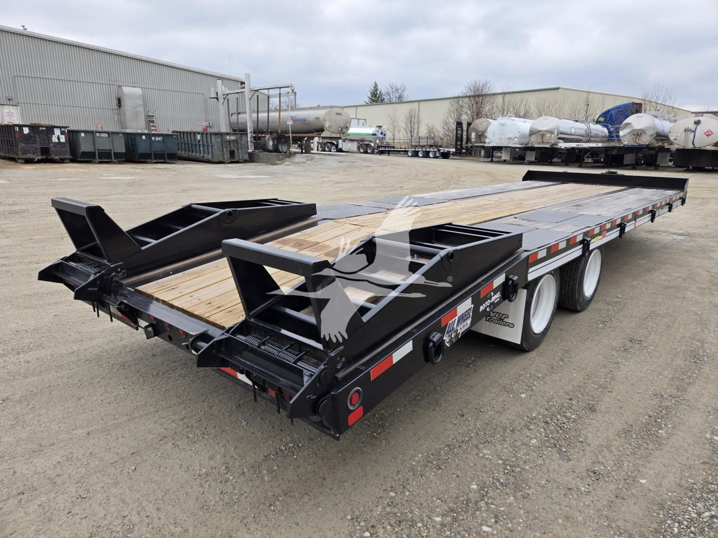 Trailer bed showing heavy-duty flatbed with black frame and dual rear ramps, wooden deck, tandem axle with white wheels, in a storage yard.