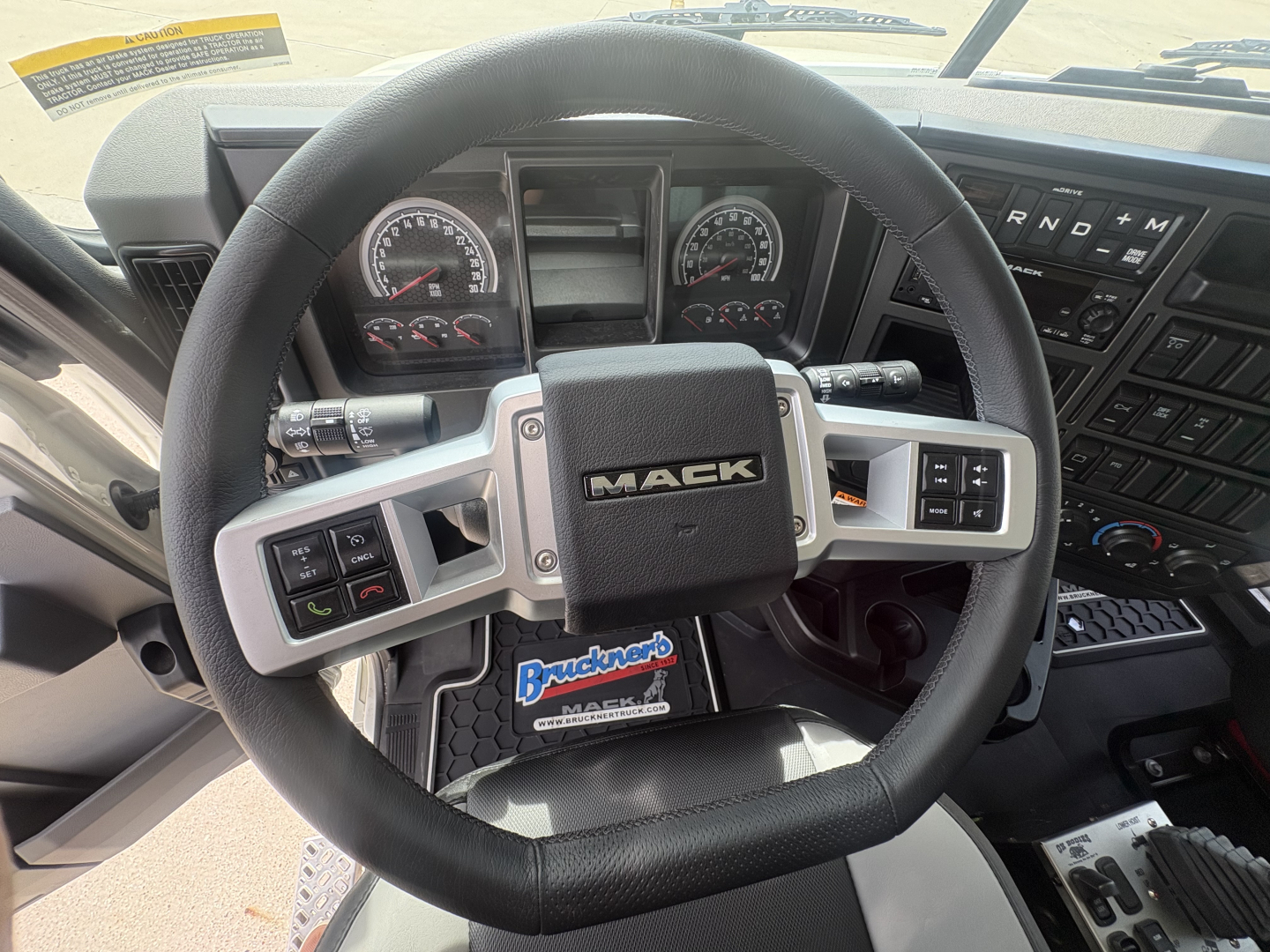 Mack truck cabin steering wheel with Mack logo, dashboard gauges, and controls visible in the dash; center console and gear shifter area.