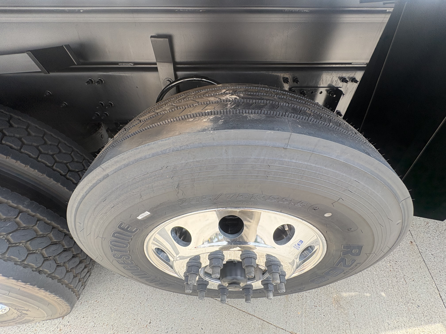 Truck wheel and dual tires showing RSTON brand wheel with lug nuts; portion of dump truck frame visible.