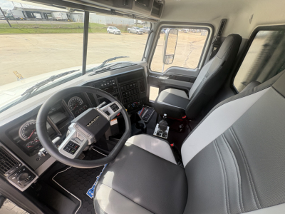 Truck interior showing steering wheel and dashboard of a new Mack GR84F cab. Visible are instrument cluster, center console with controls, gear shifter, and two-tone gray seats.