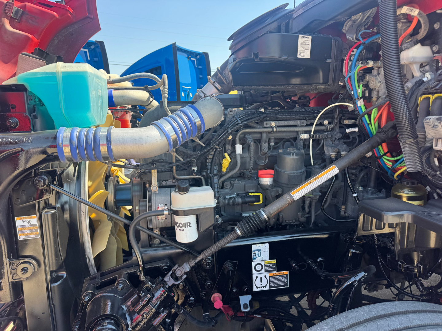 Kenworth T680 engine bay with visible hoses, battery, coolant reservoir, wiring harnesses, and air intake; various components and labels visible.