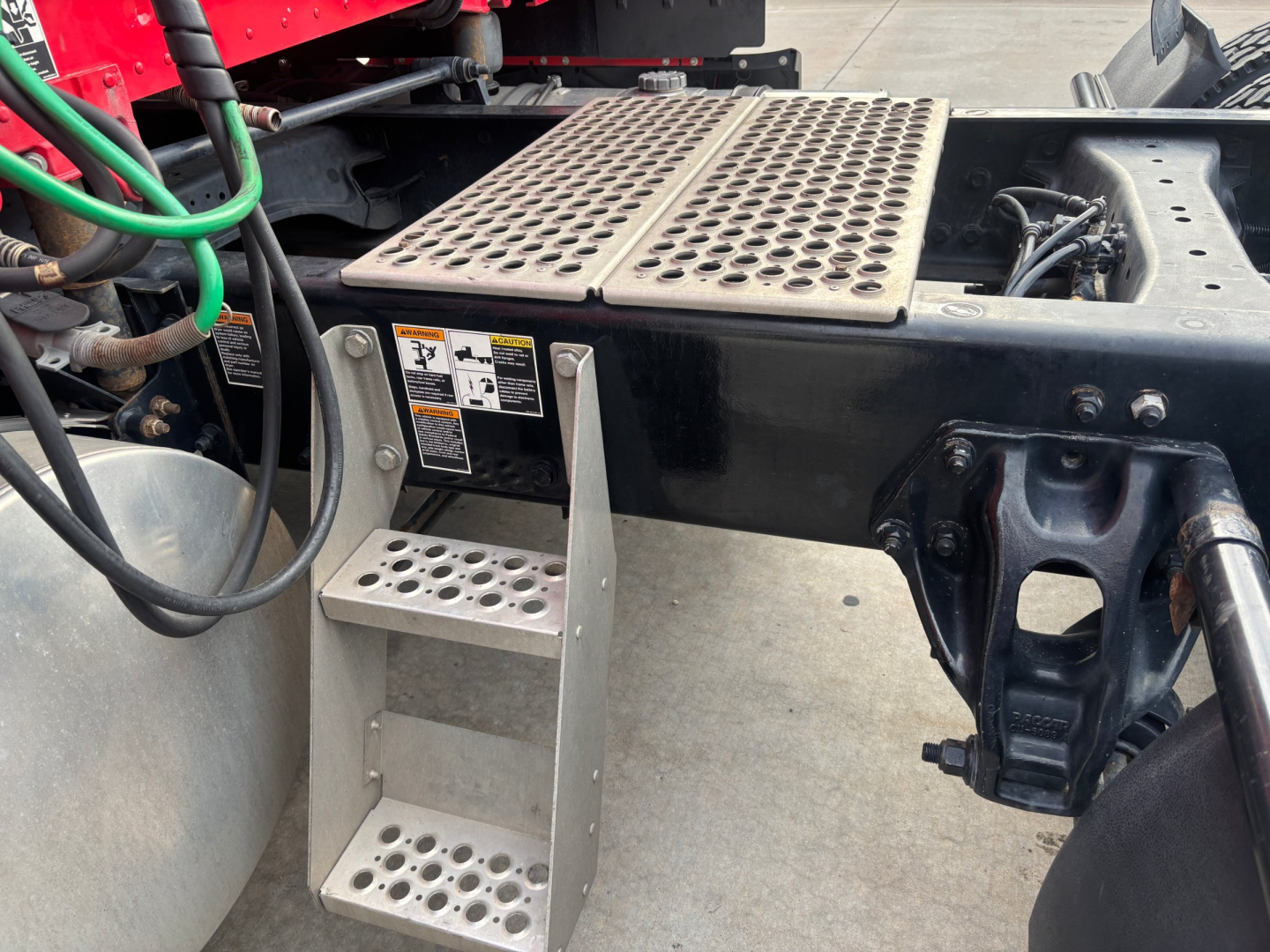 Kenworth T680 chassis step and rear cab area showing perforated metal steps and frame rails with warning labels and hydraulic lines.