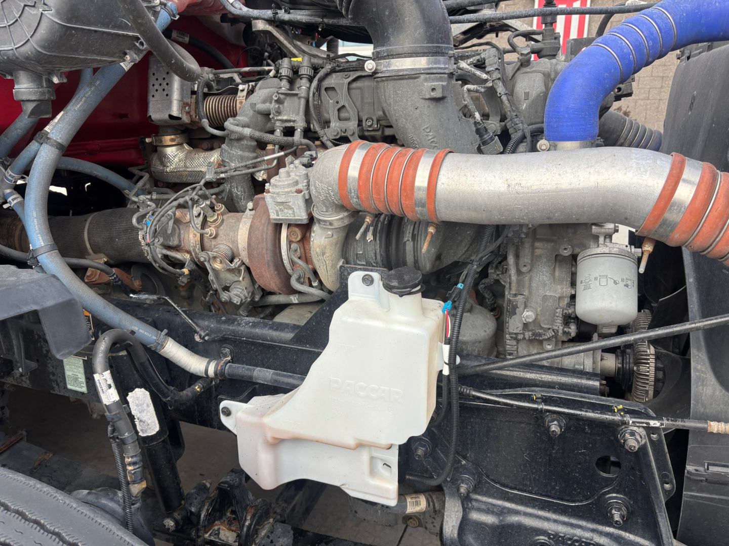 Kenworth T680 engine bay showing white coolant reservoir, turbocharger, hoses, and intake piping.
