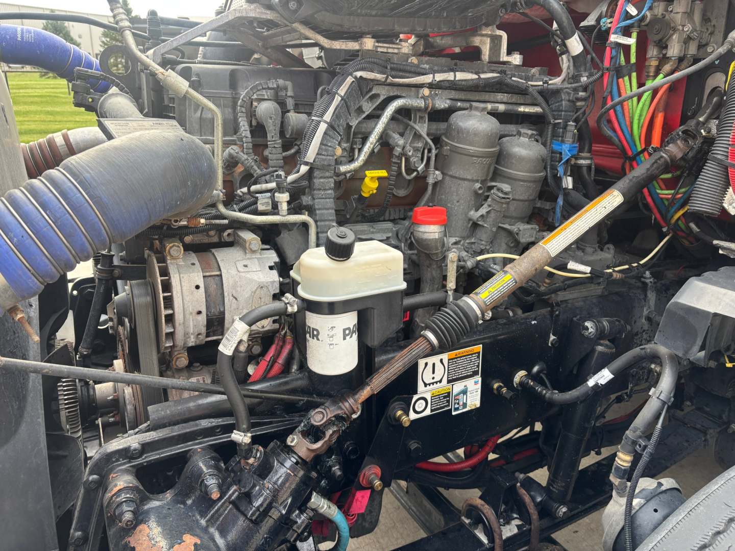 Kenworth T680 engine bay showing coolant reservoir, oil/fluids reservoirs, hoses, wiring, and turbo/intercooler area.