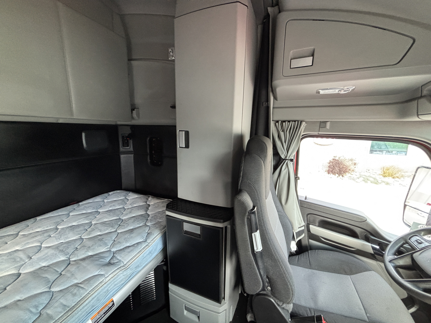 Kenworth T680 sleeper cab interior showing bunk with mattress, overhead cabinets, and driver's seat in cabin.