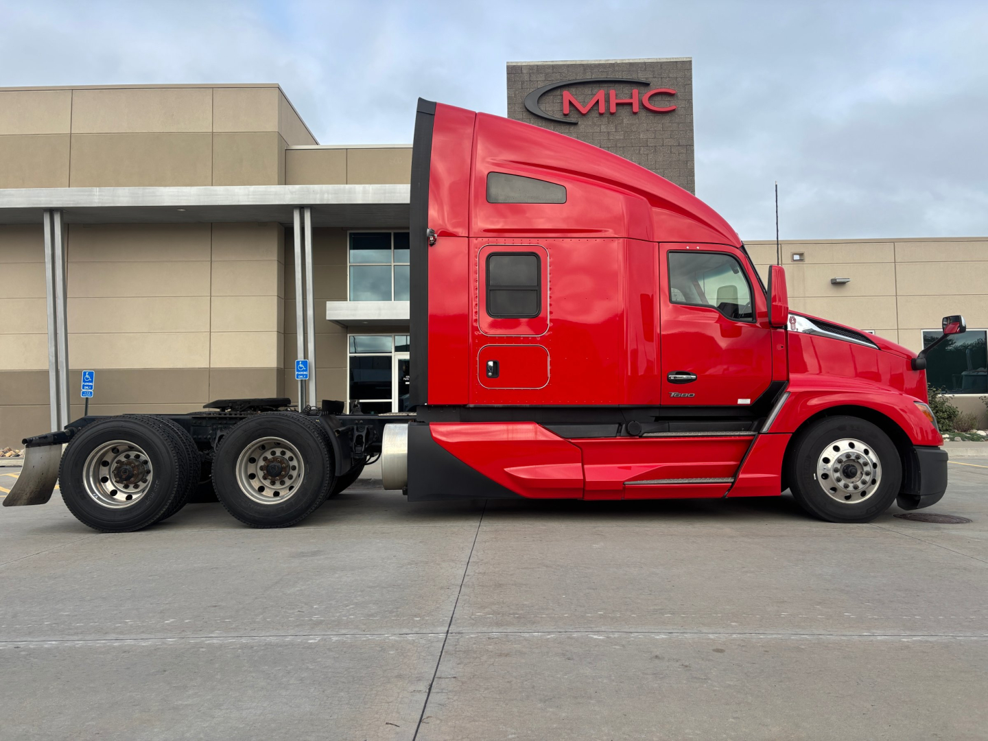 Red 2022 Kenworth T680 tractor-trailer, side profile view, sleeper cab, 6x4 configuration, parked outside dealership with MHC sign in background.