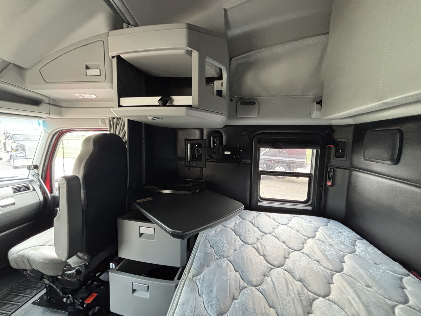 Kenworth T680 sleeper cab interior showing bunk with quilted mattress, overhead storage drawer, and compact desk area; window and driver seat visible.