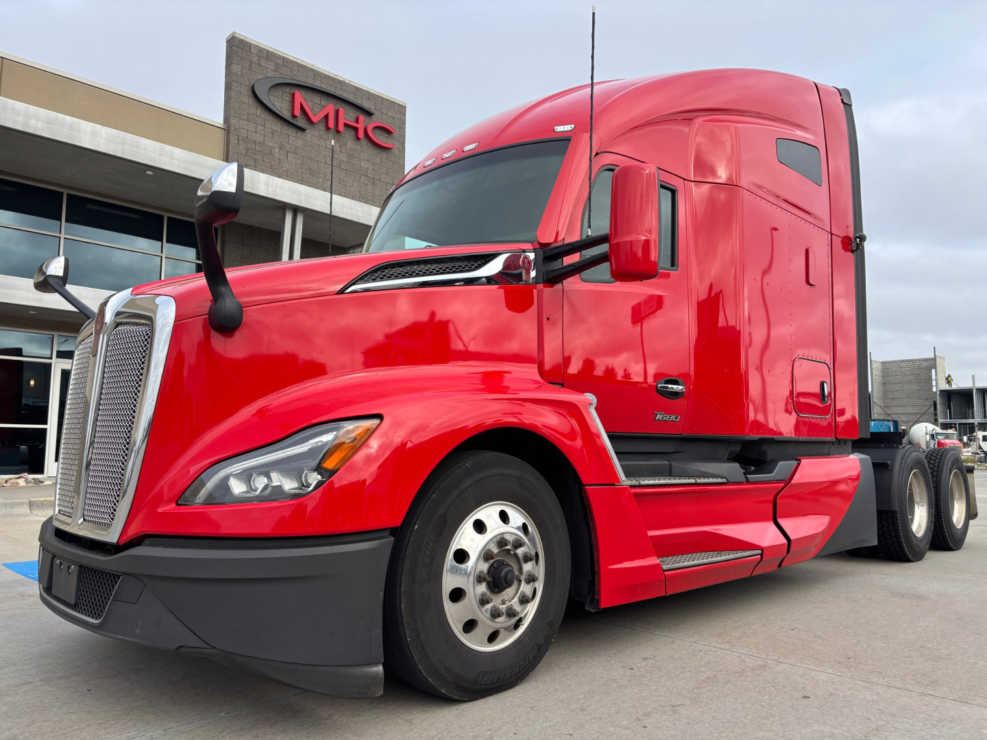 Kenworth T680 tractor truck, red exterior, front-left view, showing cab and bumper near dealership; no visible text except subtle badge near door window.