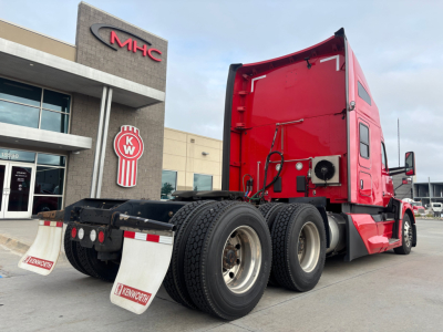Kenworth T680 truck tractor shown from rear-left side with red cab, dual rear axles, fifth wheel, and mud flaps. Building with MHC/KW logos in background.