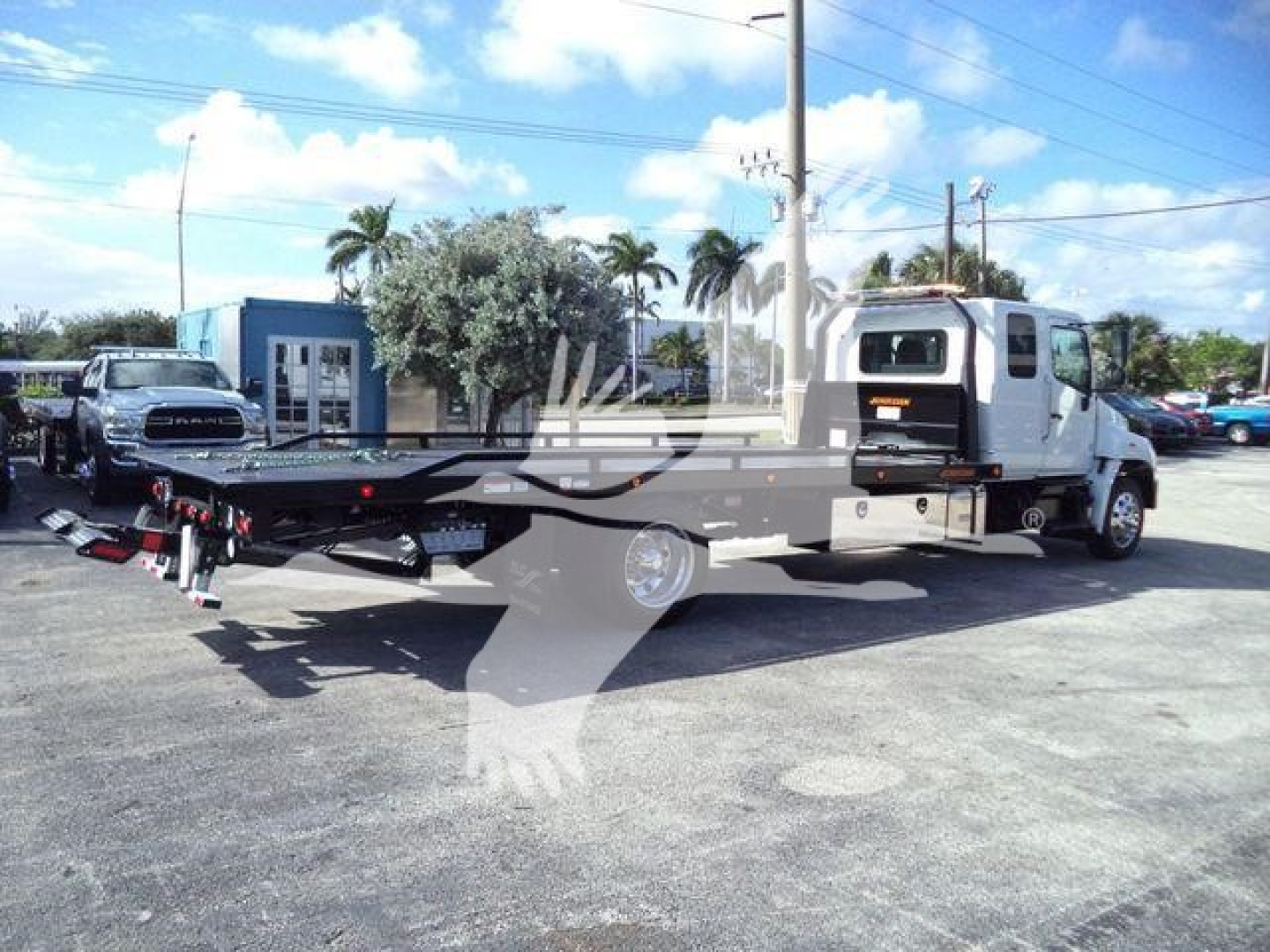 Flatbed tow truck with a white cab and a long black flatbed, parked in a lot with a blue building in background.
