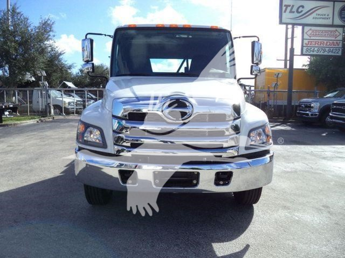 Front view of a white 2025 Hino 268A rollback truck with chrome grille and bumper, side mirrors, and cab reflection; background shows dealership lot.