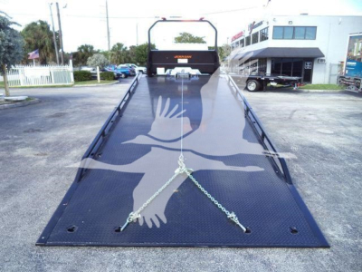 Heavy-duty Hino rollback truck flatbed with black metal ramp surface and chained anchor points, rear view showing cockpit area and dealership building in background.