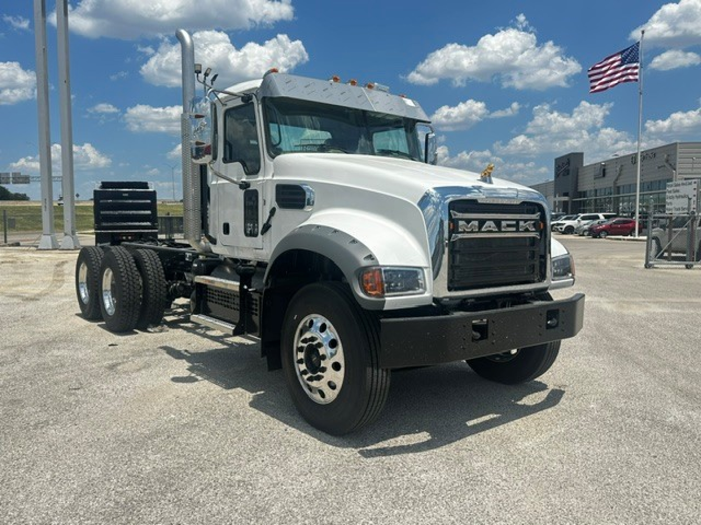 Mack GR64F cab and chassis truck side view showing front grille with MACK logo, dual rear axles, white cab, black bumper, and American flag in background.