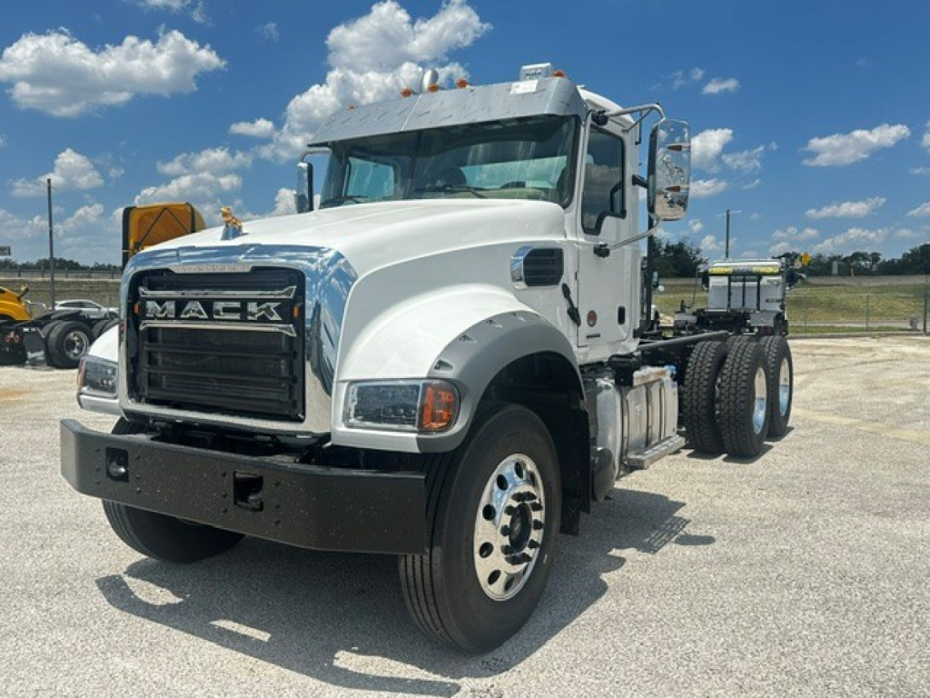Mack GR64F cab and chassis truck in white, front left view with grille MAS K letters, large side mirrors, dual rear axles, chrome bumper, clear blue sky.