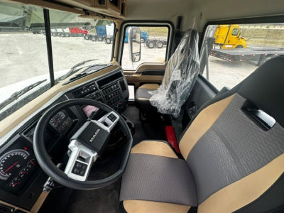 Cab interior of a 2027 Mack GR64F showing steering wheel, dashboard, driver seat with plastic cover, and passenger seat with two-tone upholstery.