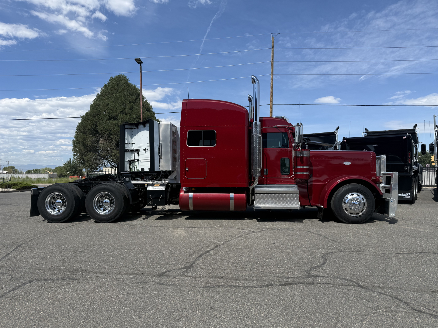 Peterbilt 389 conventional sleeper truck in red, side view showing cab, chrome fuel tanks, and dual rear axles. No visible text on the truck components in the photo.