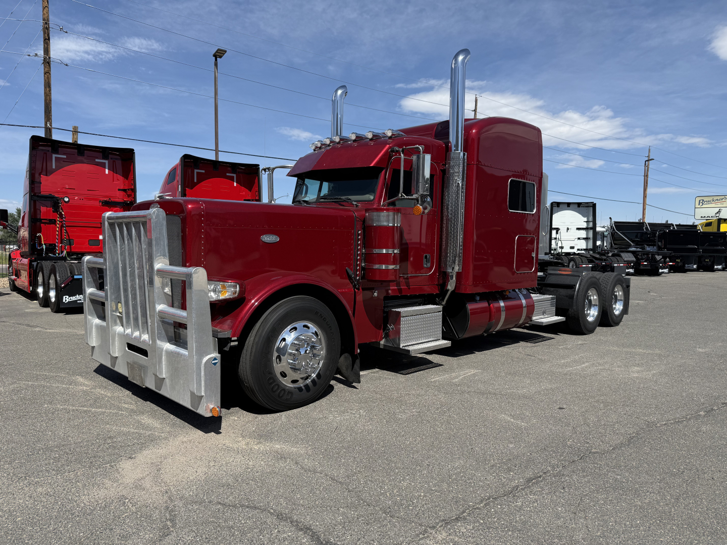 Peterbilt 389 conventional sleeper truck in red with chrome exhaust stacks and a front bumper guard visible in a dealership lot.