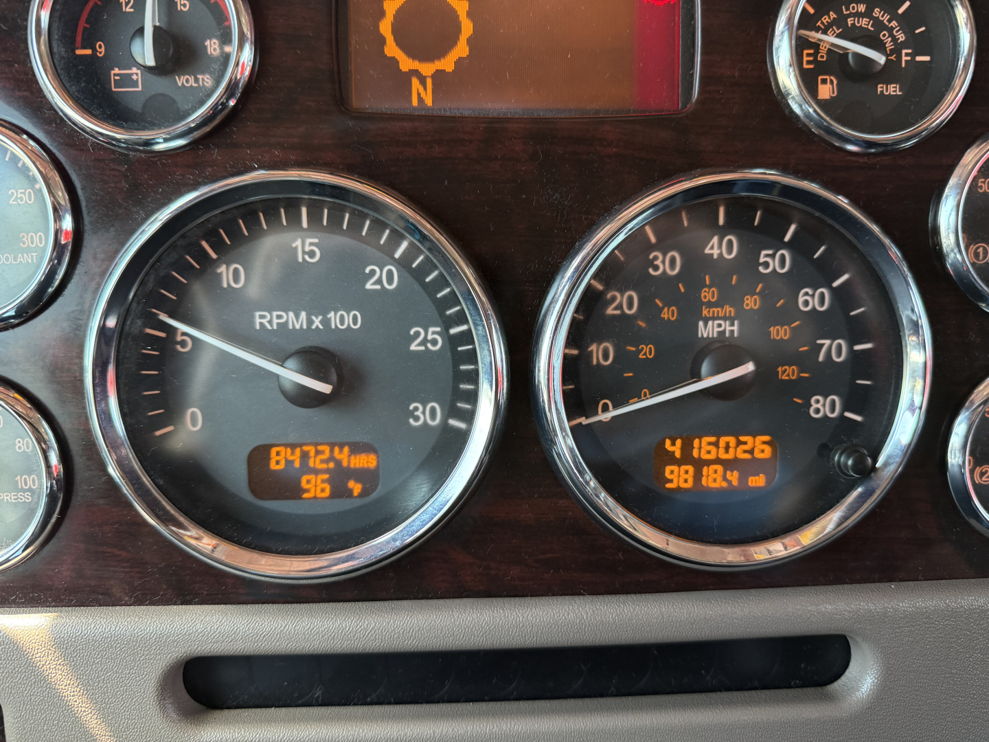 Peterbilt 389 dash cluster showing RPM x1000 gauge at 0-30, speedometer, gear indicator, and digital readouts; focus on gauges and orange warning icon.